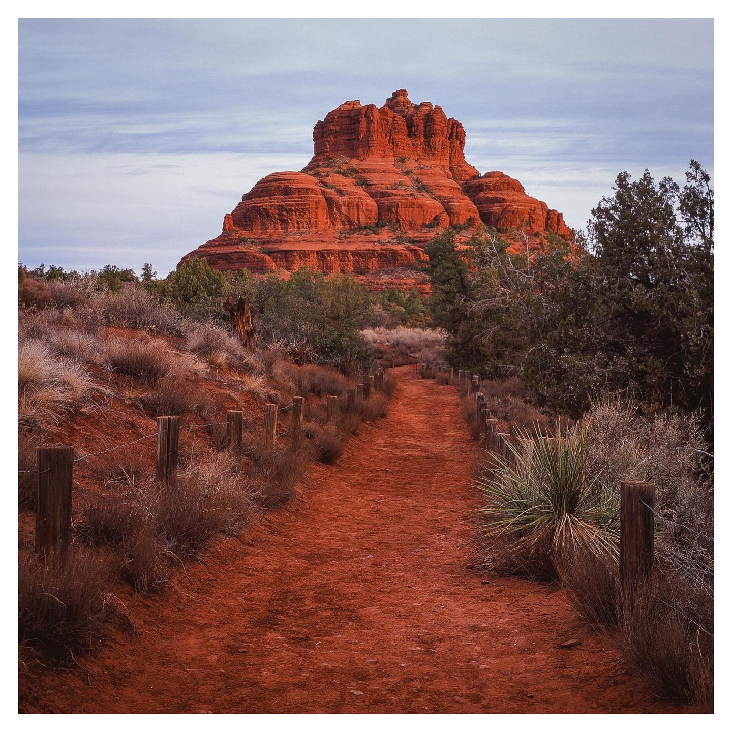 A red dirt path leads towards a large red rock formation under a cloudy sky.