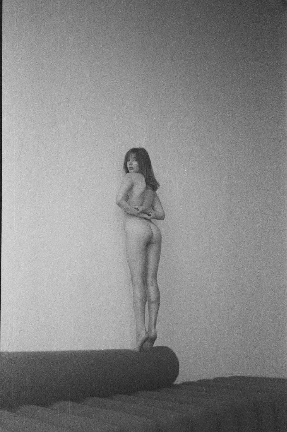 A nude woman stands on her toes on a cylindrical object, looking over her shoulder at the viewer.