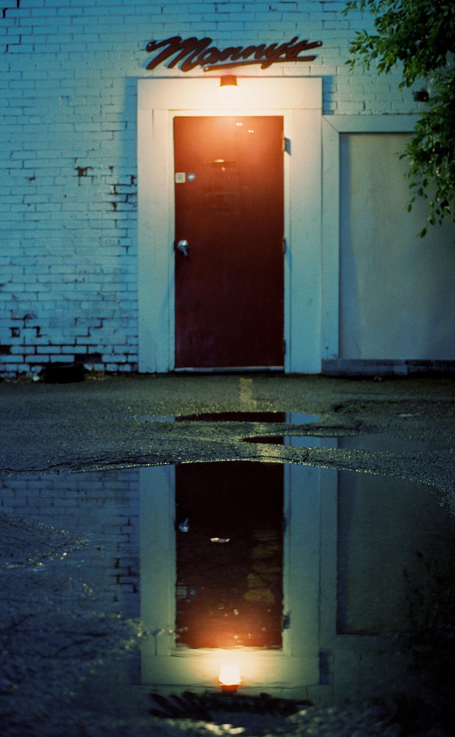 A red door with a light above it and a sign that reads "Manny's" is reflected in a puddle on the ground.
