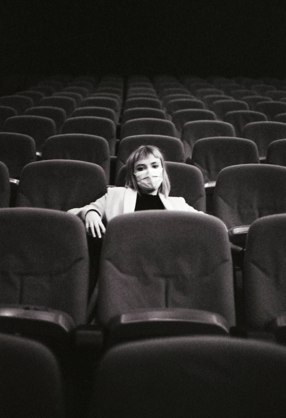 A woman wearing a face mask sits alone in a movie theater with many empty seats.