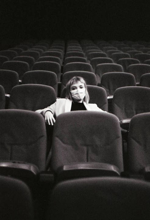 A woman wearing a face mask sits alone in a movie theater with many empty seats.