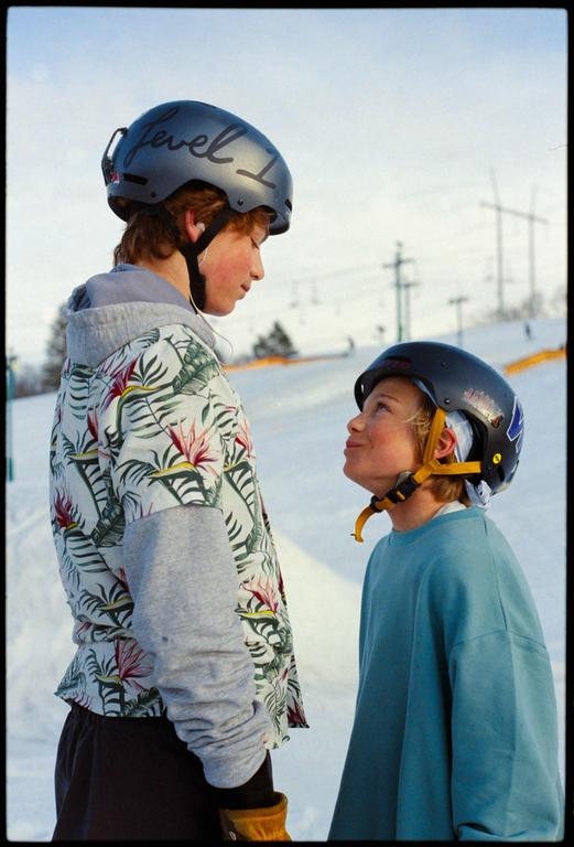 Two boys wearing helmets stand on a snowy slope, looking at each other.