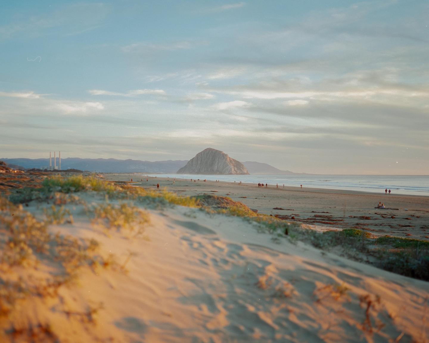 A wide sandy beach with people walking along the shore is visible, with a large rock formation and mountains in the distance under a cloudy sky.
