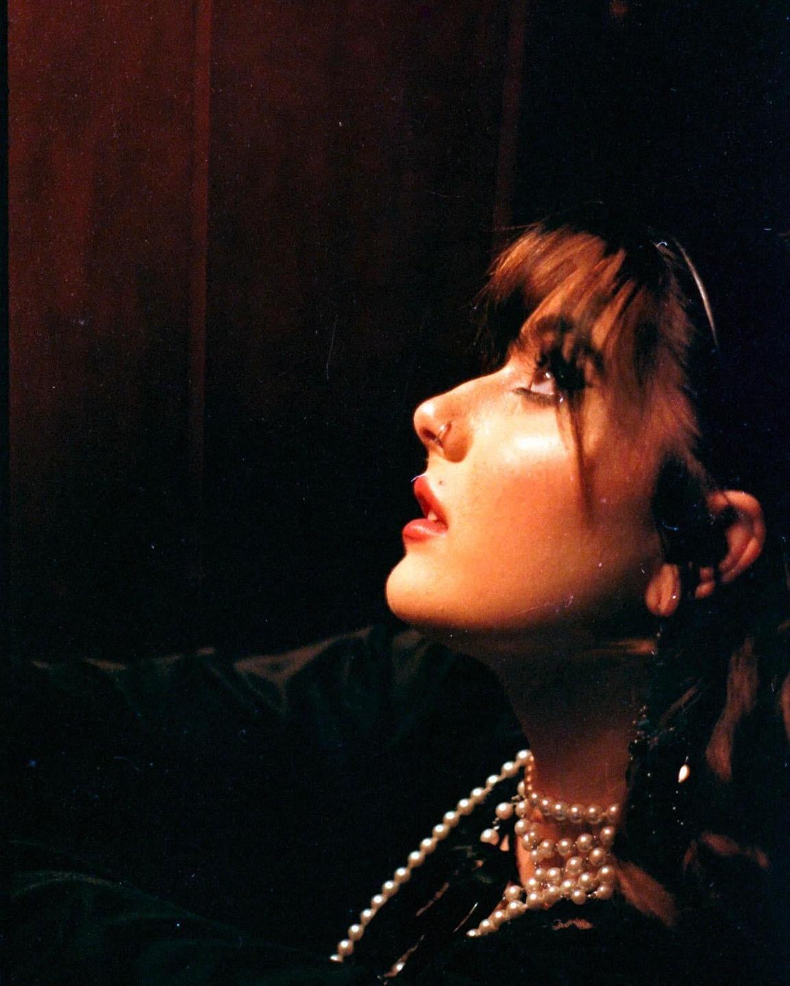 A woman with bangs and a pearl necklace looks up, illuminated by a light source from above.