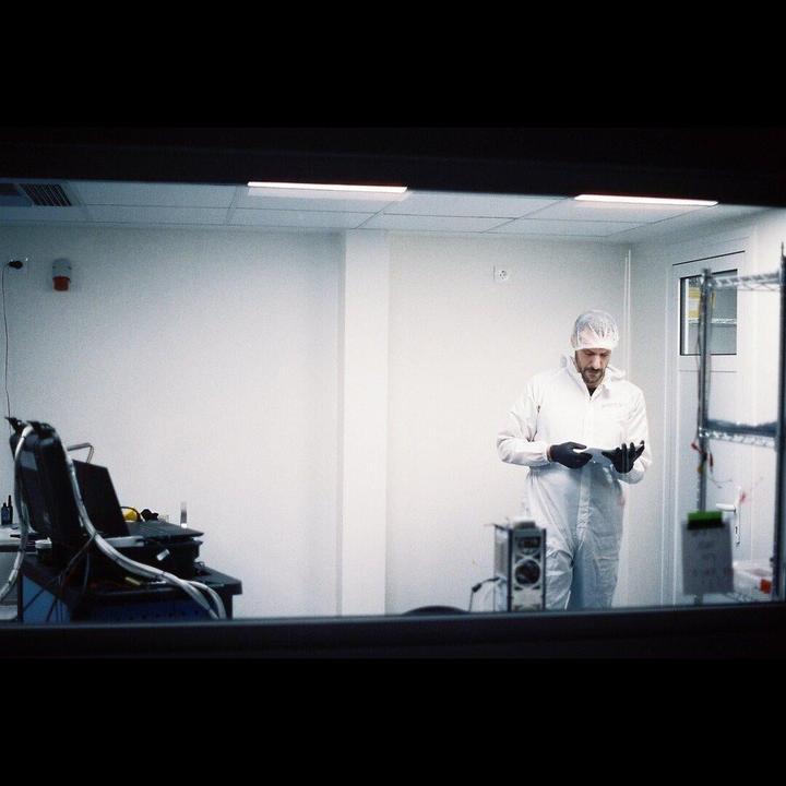 A man in a cleanroom suit and hairnet stands in a white room, looking down at a tablet in his gloved hands.