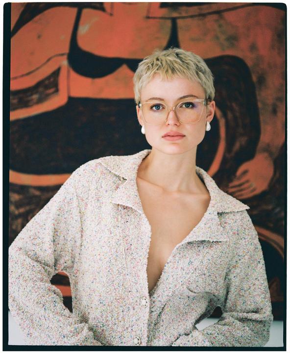 A woman with short blonde hair and glasses wears a textured jacket and pearl earrings, standing in front of an abstract painting.