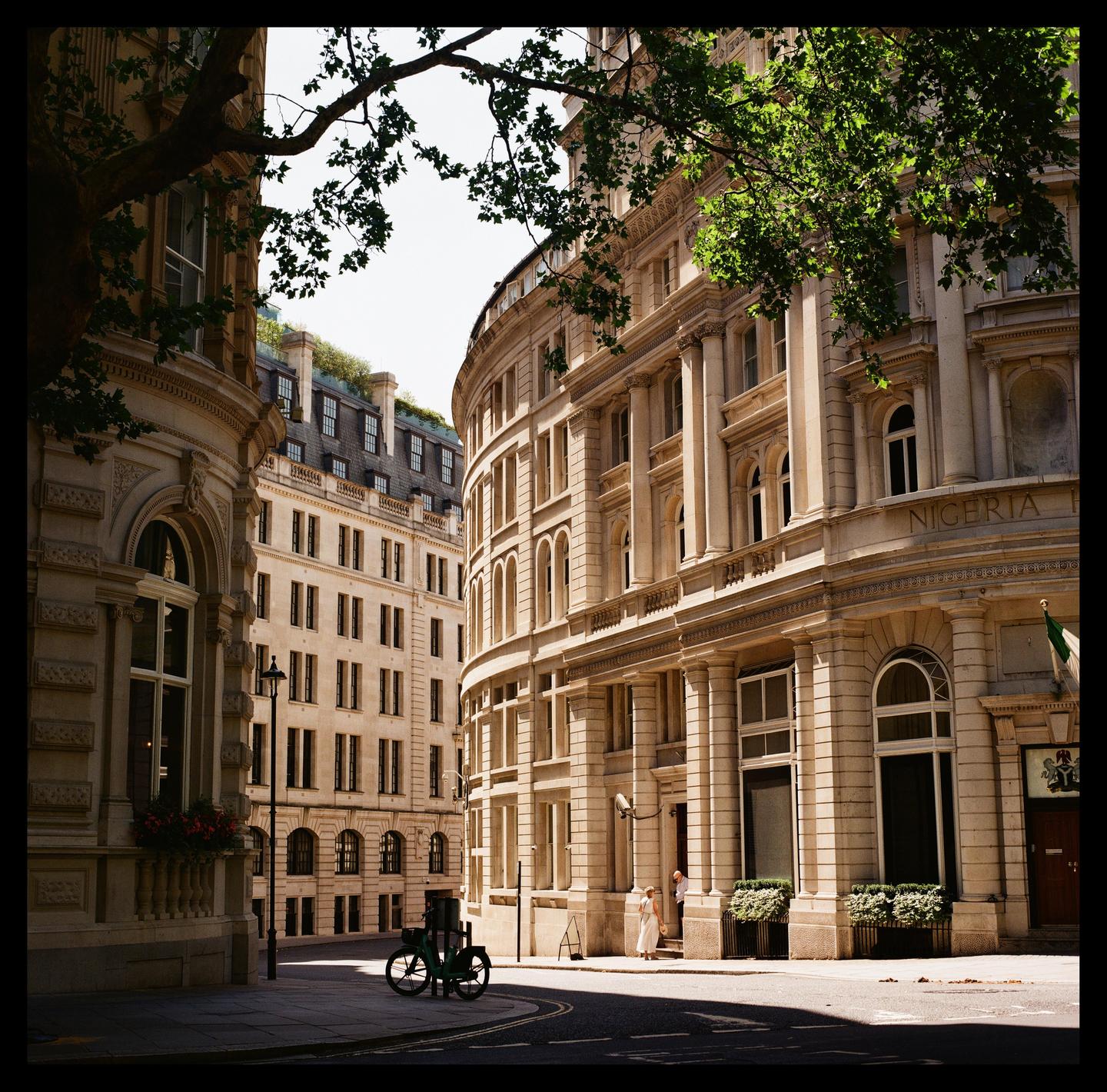 A street in London is lined with ornate buildings, with a bicycle parked on the corner and two people standing near a doorway.