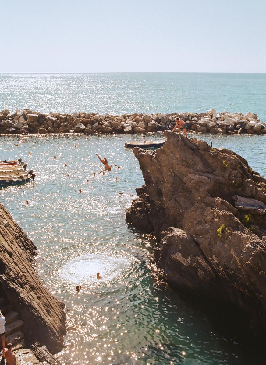 A person dives into the sparkling water from a rocky cliff, while other people swim and stand on nearby rocks.