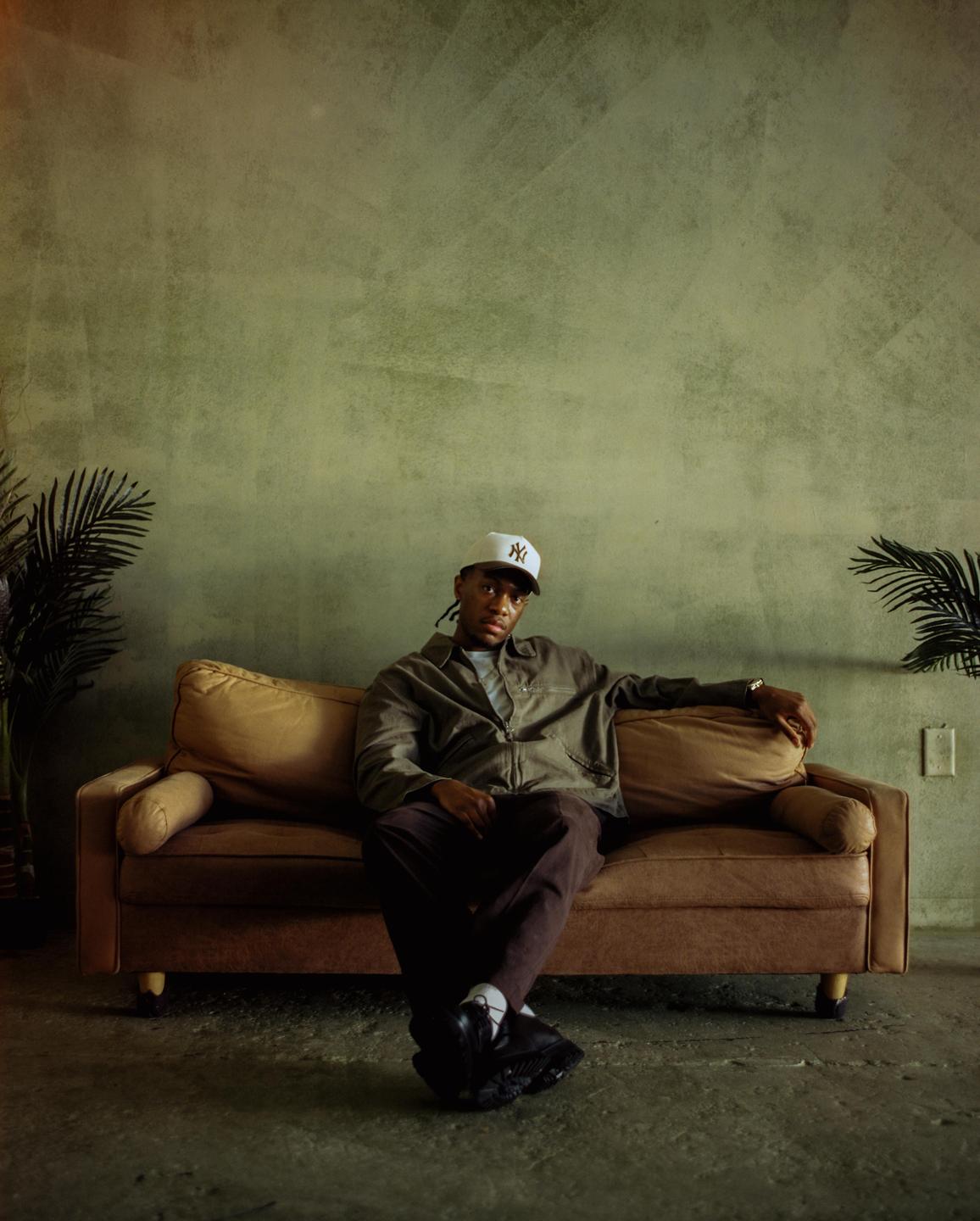 A man wearing a baseball cap and jacket sits on a brown sofa with plants on either side.