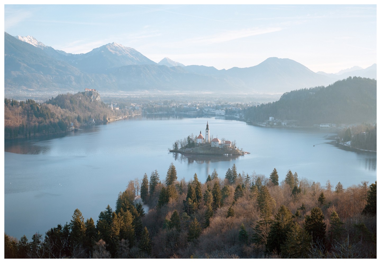 A small island with a church and buildings sits in the middle of a lake surrounded by mountains and a town.