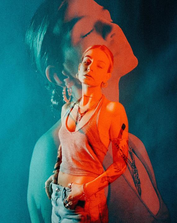 A woman with tattoos and jewelry is shown in a double exposure with a second, larger portrait of a woman's head in the background.