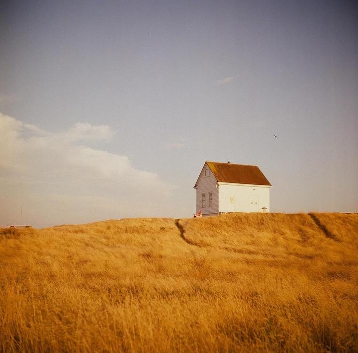 A white house with a brown roof sits atop a hill covered in golden grass under a partly cloudy sky.