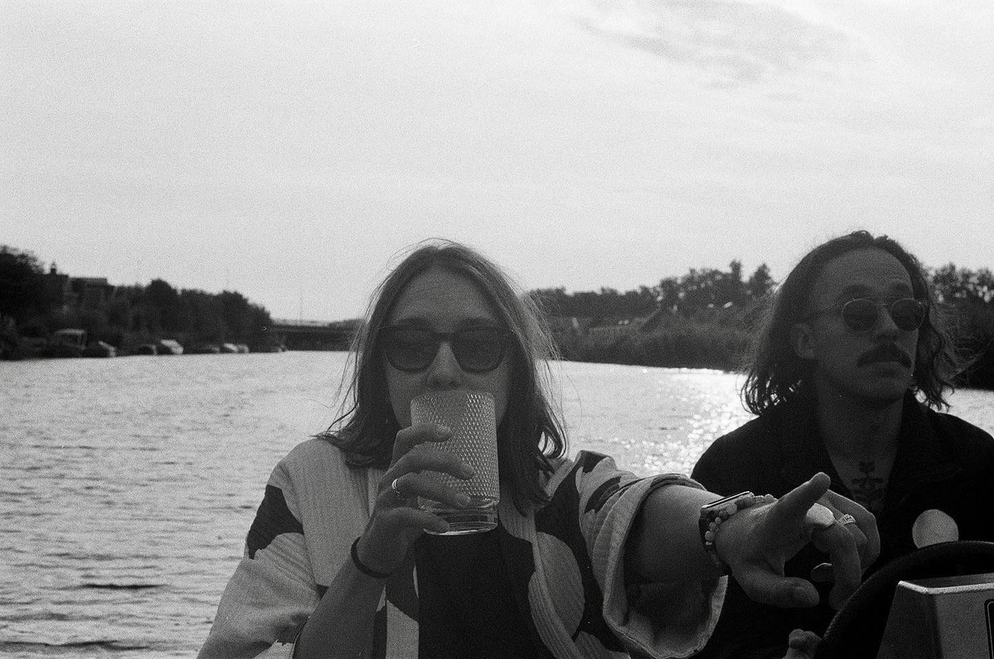 A woman drinks from a glass while a man points forward on a boat.