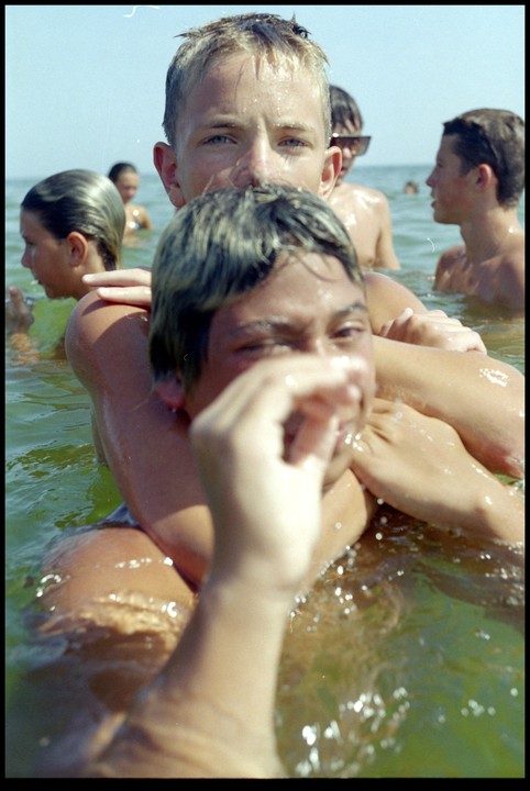 A boy with wet hair looks directly at the camera while another boy's head and arms are in the foreground, partially obscuring him, with other people swimming in the background.