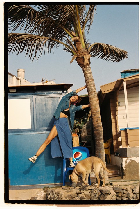 A woman leans against a palm tree while a dog drinks from a bowl next to a blue chair.