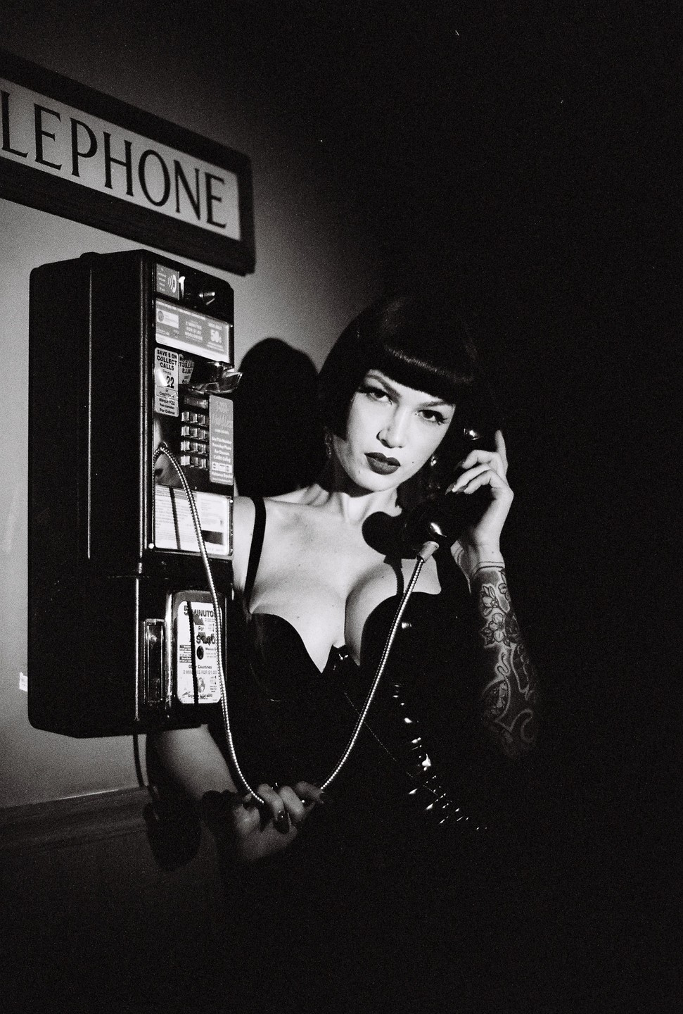 A woman in a black top holds a telephone receiver to her ear while standing next to a payphone.