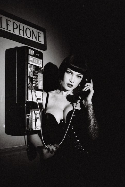 A woman in a black top holds a telephone receiver to her ear while standing next to a payphone.