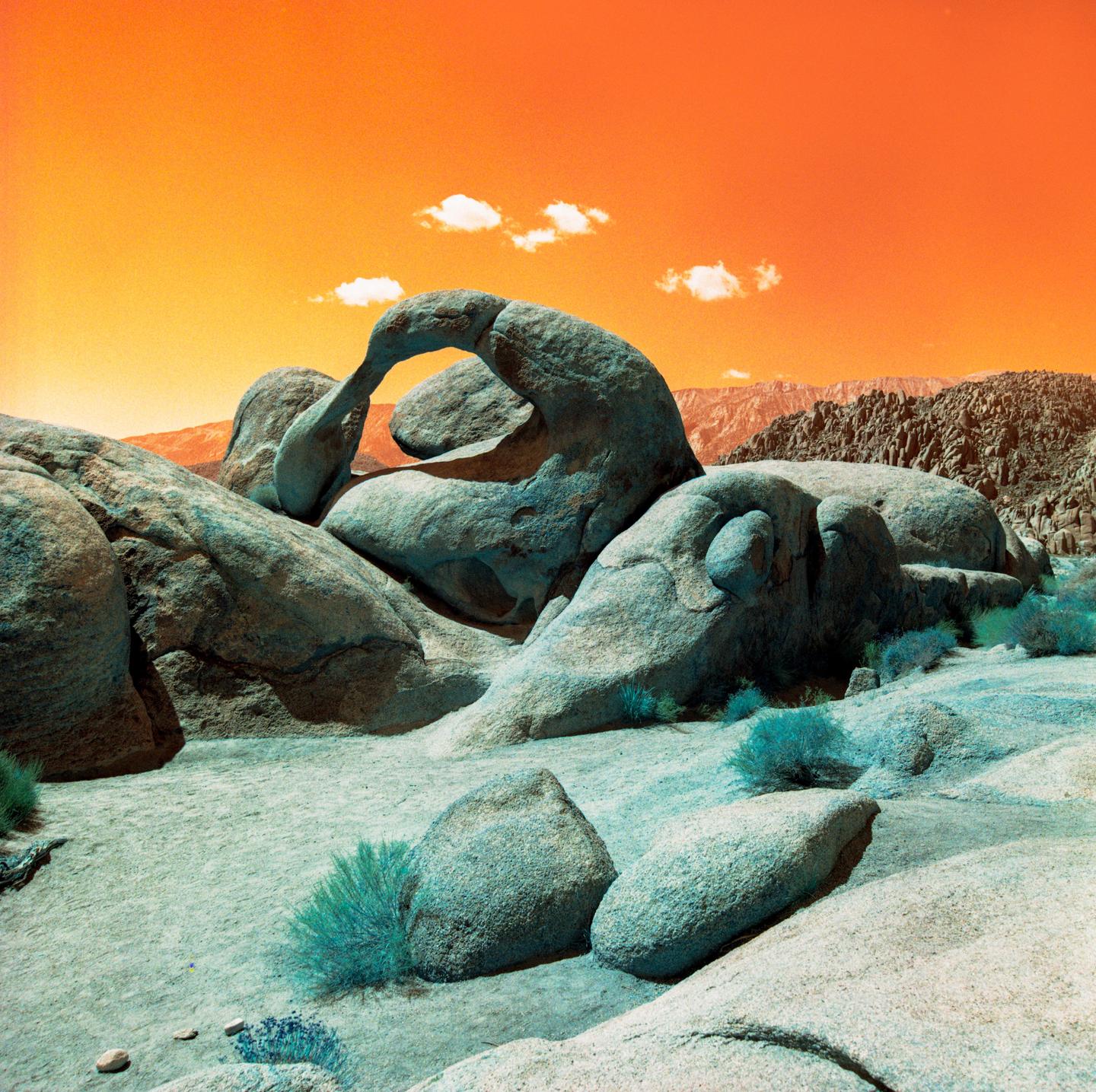 A large rock formation with an arch is visible under an orange sky with two small clouds.