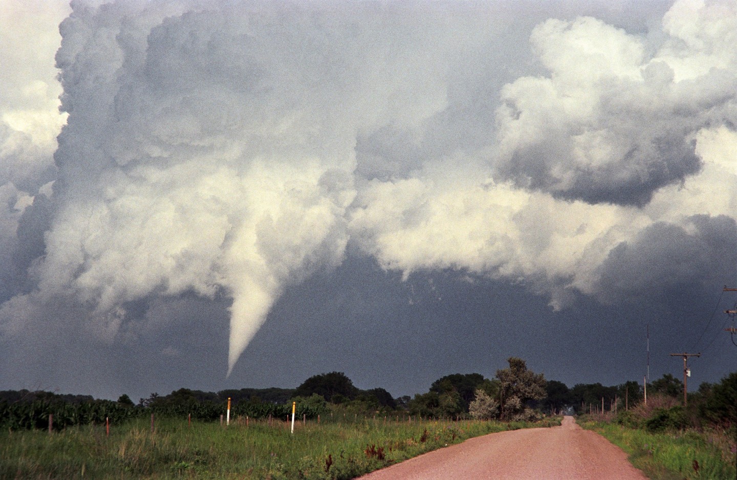 A tornado descends from dark storm clouds over a rural landscape with a dirt road in the foreground.