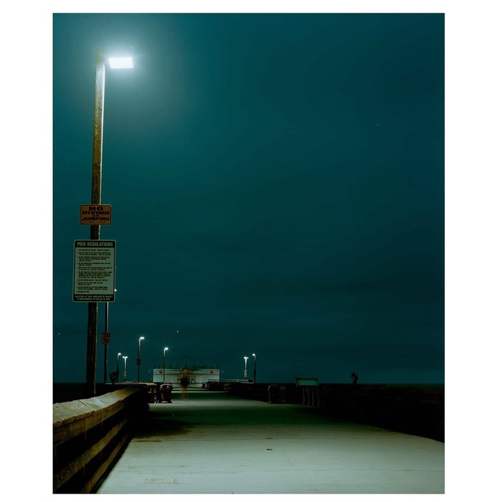A brightly lit streetlamp stands on a pier with regulations signs under a dark night sky.