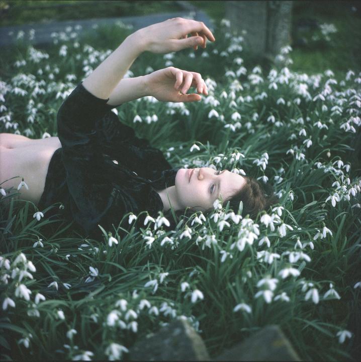 A woman with dark hair lies in a field of white snowdrop flowers, looking upwards with her arms raised.