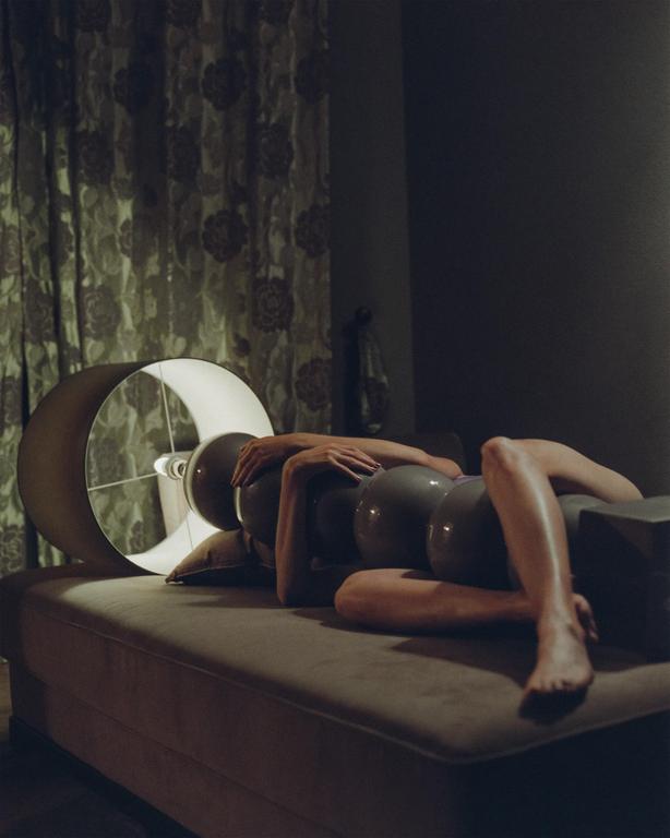 A woman lies on a sofa with balloons on her body and a lamp behind her.