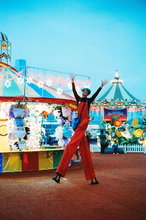 A person on stilts with arms raised stands in front of a carnival game booth with stuffed animals and a carousel in the background.