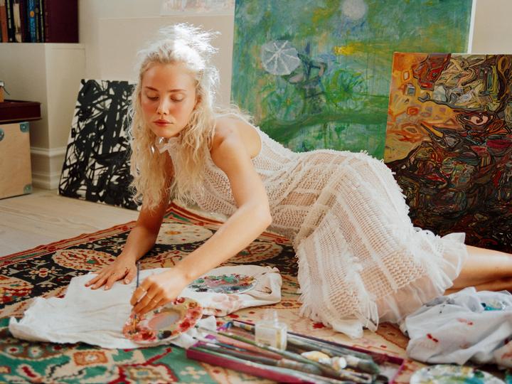 A woman lies on a patterned rug, painting on a piece of fabric with a palette and brushes beside her.