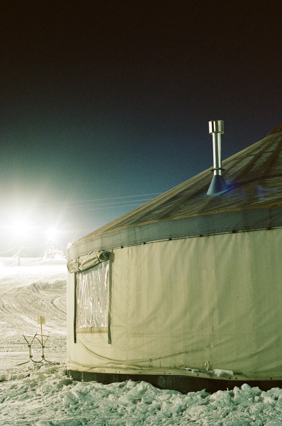 A yurt with a chimney on its roof stands in a snowy landscape at night, illuminated by a bright light in the distance.