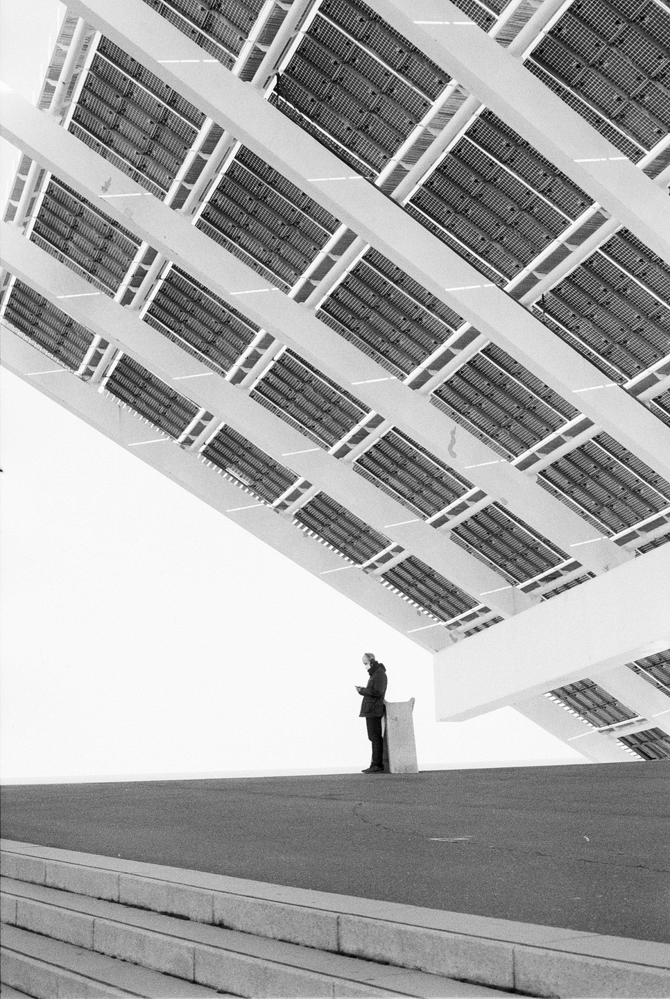 A man stands on a paved surface under a large, angled structure with many rectangular panels.