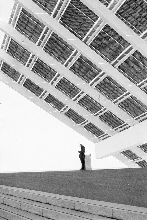 A man stands on a paved surface under a large, angled structure with many rectangular panels.