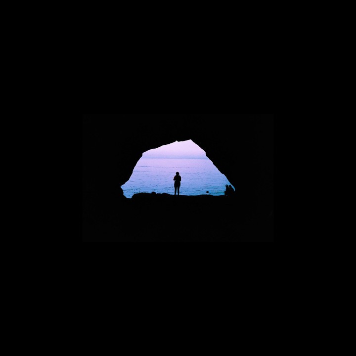 A person stands silhouetted in a cave opening looking out at the ocean under a purple sky.