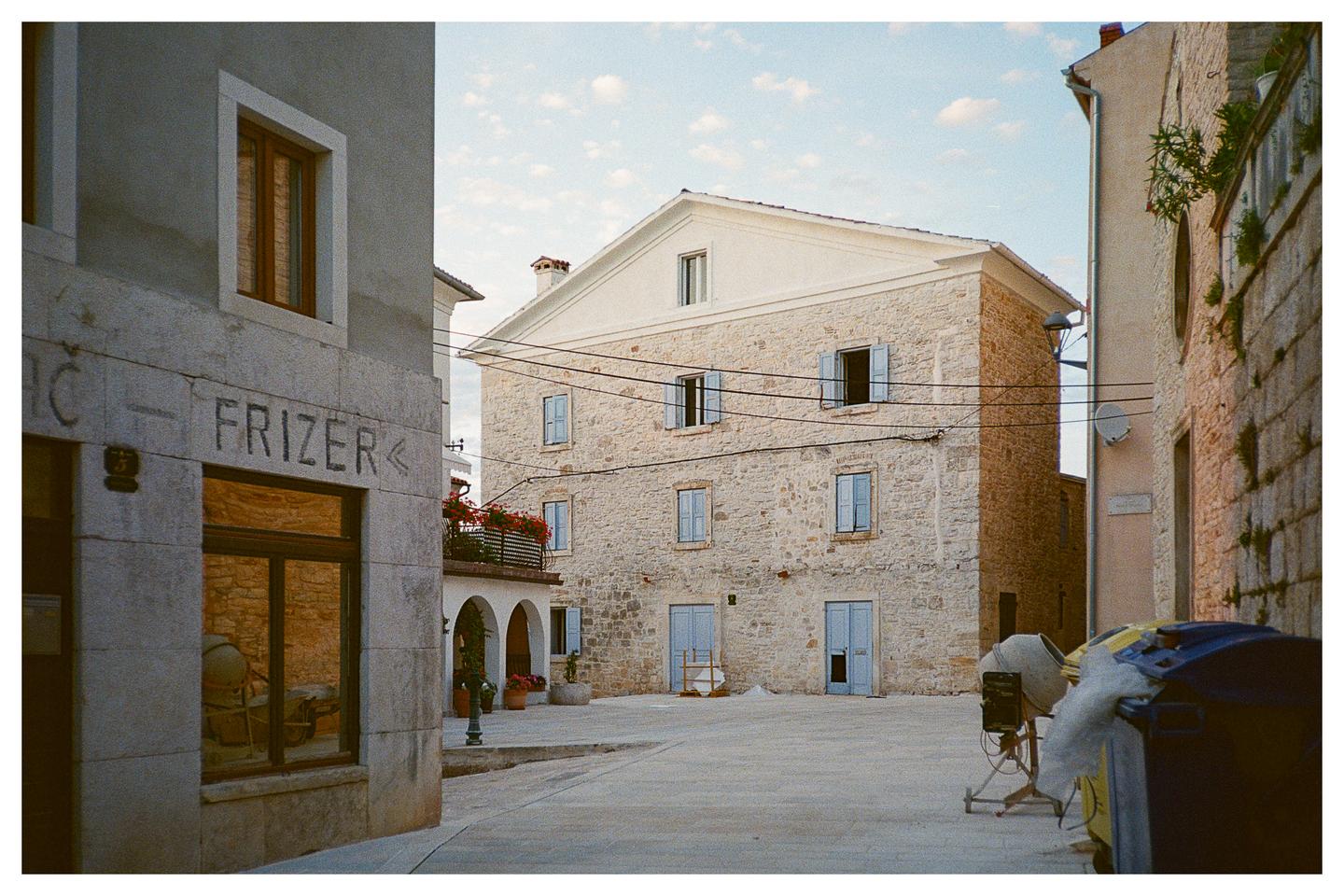 A stone building with blue shutters and a balcony with red flowers stands next to a building with a "FRIZER" sign.
