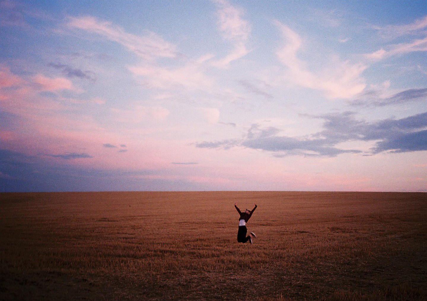 A woman jumps in a field under a pink and blue sky.