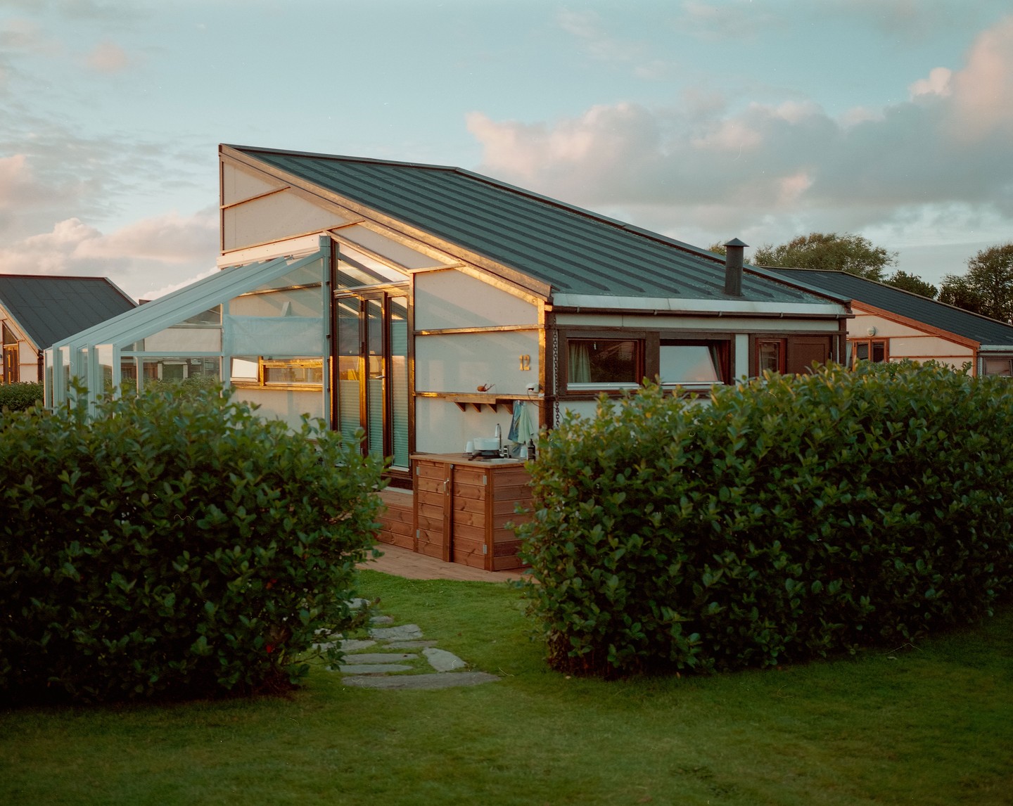 A modern house with a greenhouse extension is seen from a grassy yard with hedges.