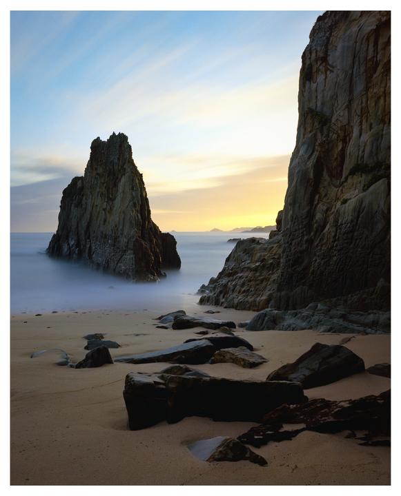 Two large rock formations stand on a sandy beach with the ocean and a colorful sky in the background.