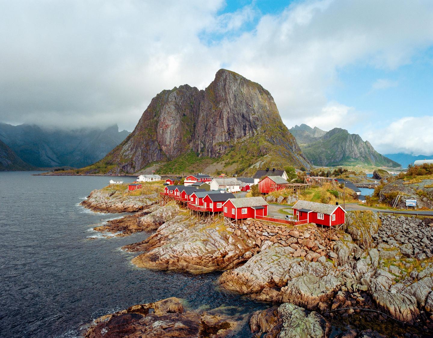 Red houses are built on a rocky coastline with a large mountain in the background.