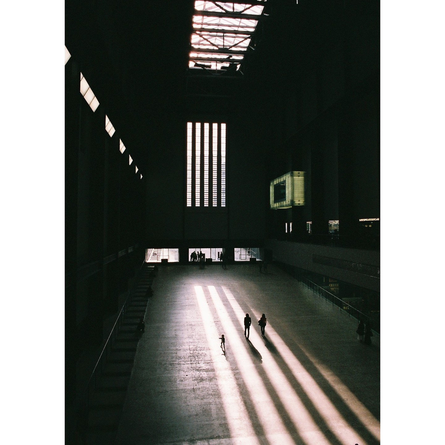 Three people walk across a large, dark hall illuminated by strong diagonal shafts of sunlight from a high window.