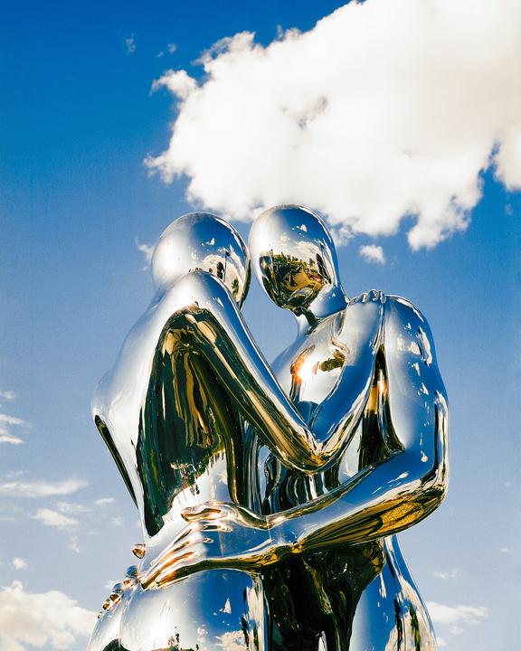 Two chrome statues of people embracing stand against a bright blue sky with a large white cloud.