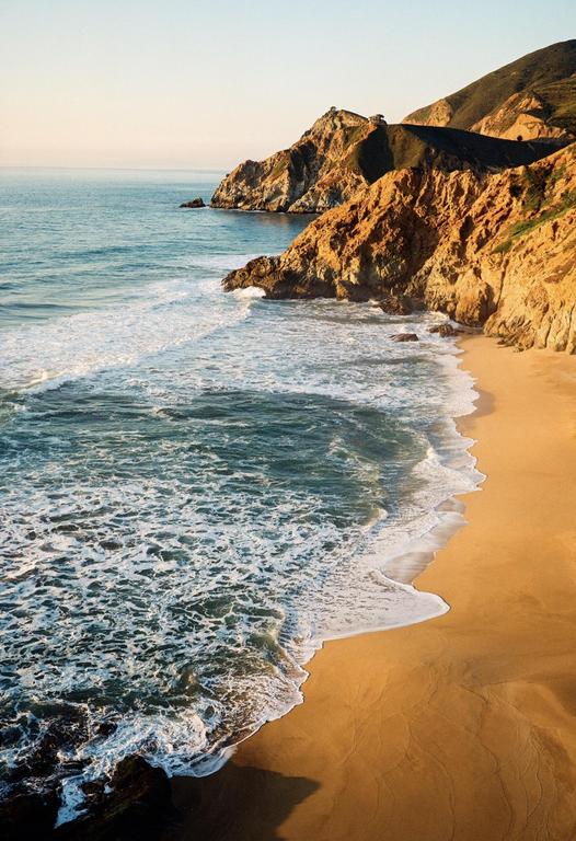 Waves crash on a sandy beach next to a rocky cliff face under a clear sky.