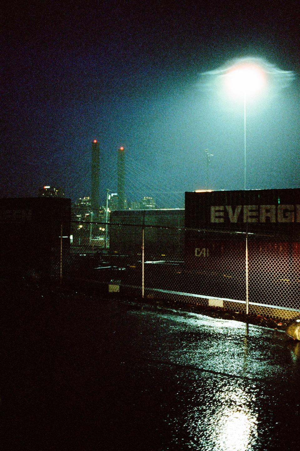 A wet street at night shows a chain link fence, shipping containers, and industrial structures in the distance.