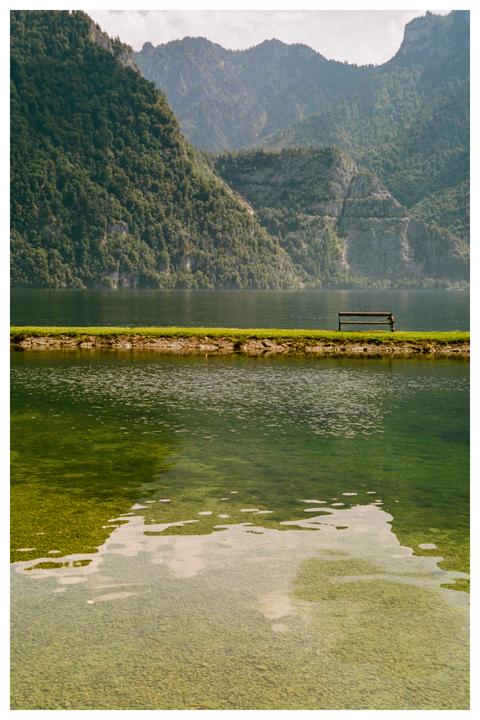 A wooden bench sits on a narrow strip of land between two bodies of water, with mountains in the background.