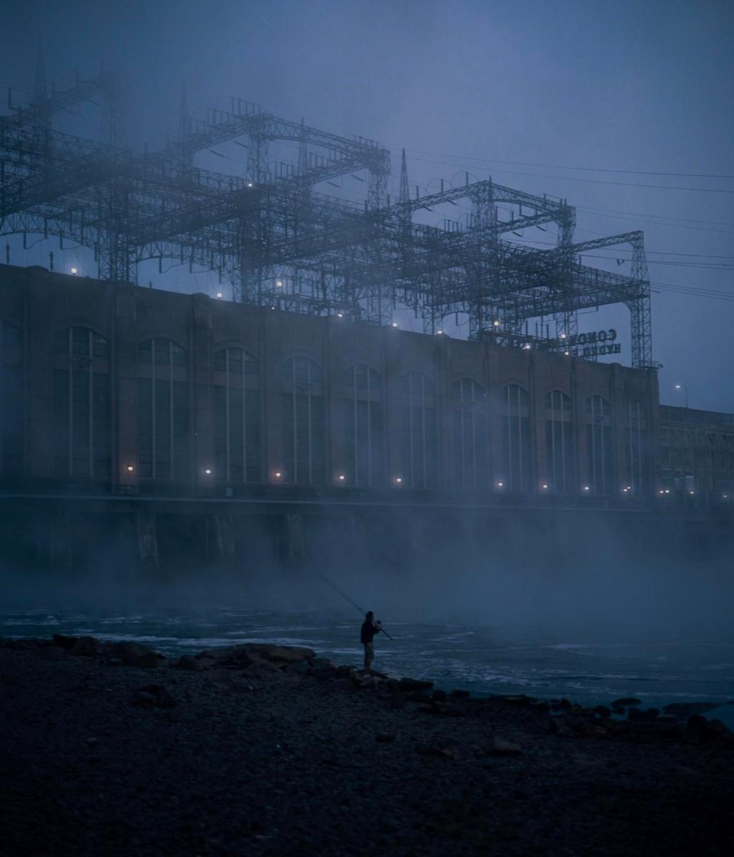 A person stands on a rocky shore fishing in front of a large building with power lines and mist.
