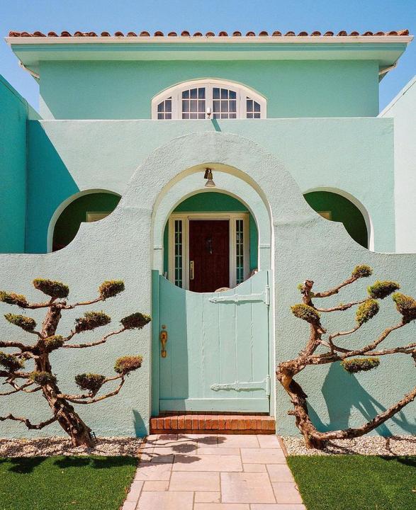 A light blue house with a dark wood door and a light blue gate is framed by two sculpted trees.
