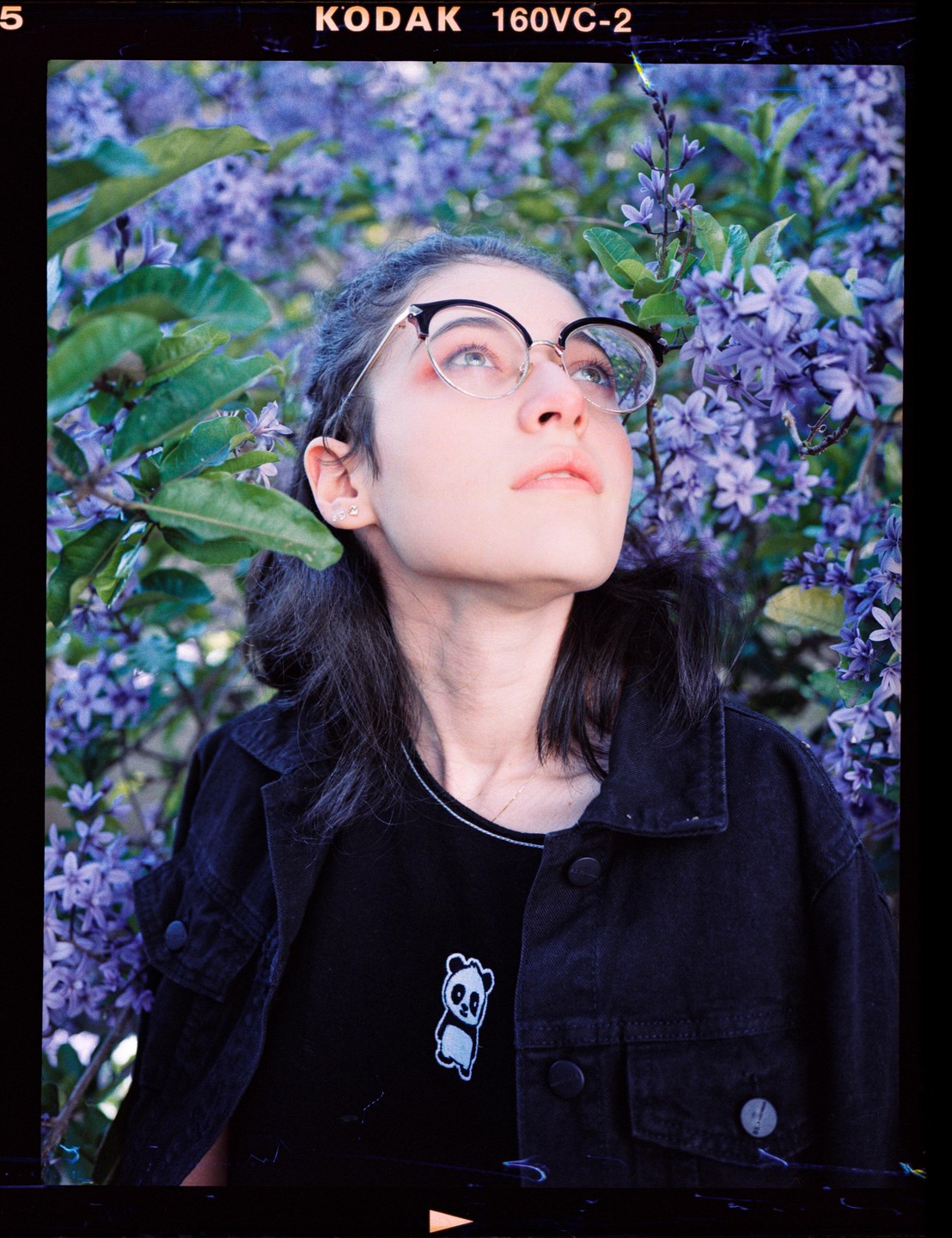 A woman with glasses looks up, surrounded by purple flowers and green leaves.