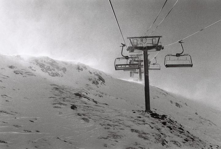 A ski lift ascends a snow-covered mountain with blowing snow.