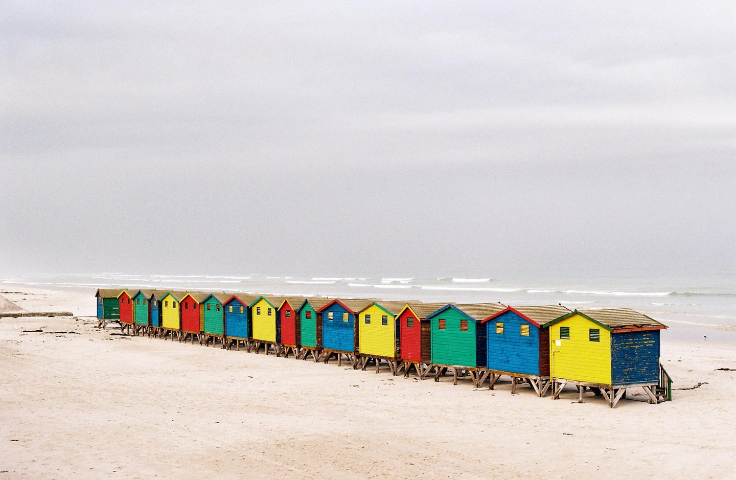 A long row of colorful beach huts stands on a sandy beach under a gloomy sky with ocean waves in the background.