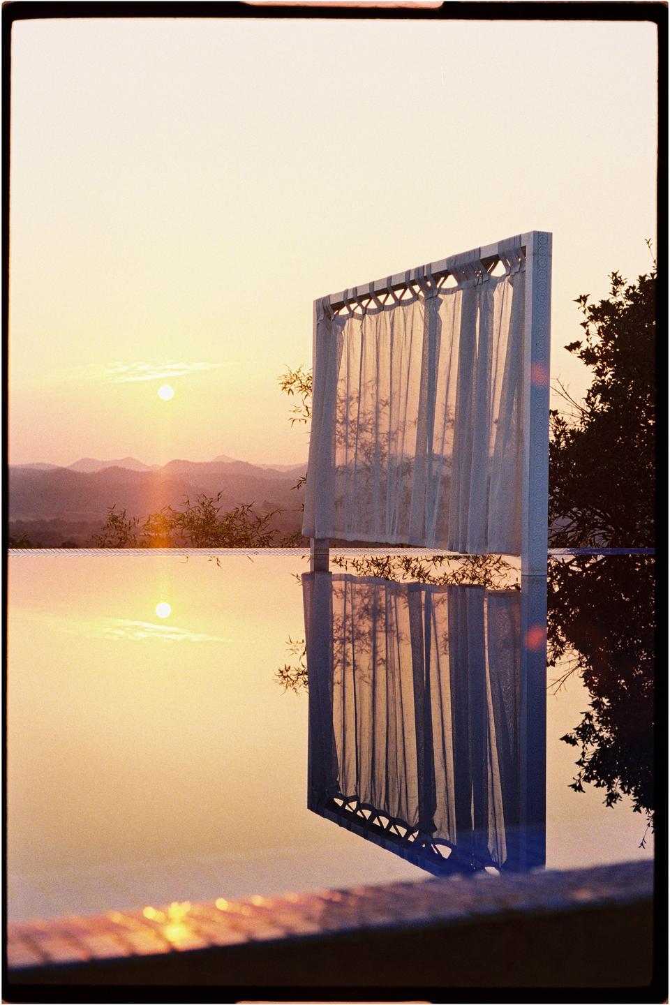 A sunset over mountains is reflected in a swimming pool next to a curtained structure.