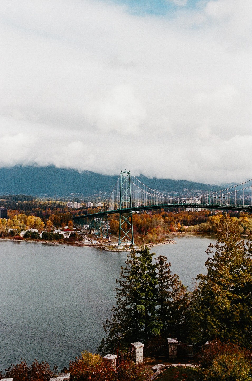 A suspension bridge spans a body of water with autumn trees and mountains in the background under a cloudy sky.