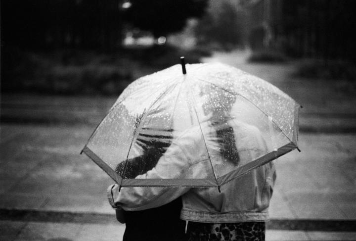 A couple stands under a clear umbrella covered in raindrops, seen from behind.